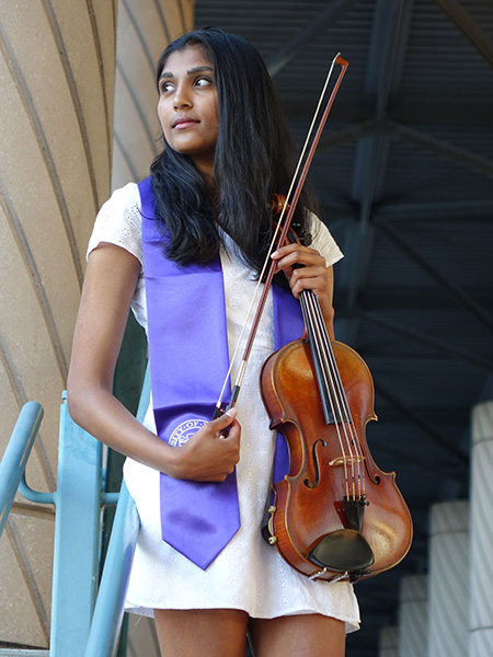 Meghna Shankar, wearing a purple sash and white dress, holds a viola in her left hand and the bow in her right. She is looking to her right while standing in front of a blue railing. 