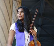 Meghna Shankar, wearing a purple sash and white dress, holds a viola and bow while looking to her right. She is standing in front of a brown and black background.
