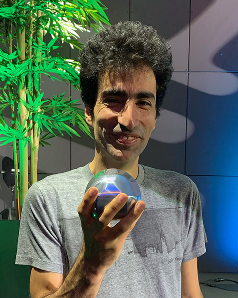 Shayan Oveis Gharan, clad in a grey t-shirt and standing in front of a tall potted tree, holds up a shiny sphere-like object in one hand while smiling for the camera.