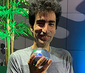 Shayan Oveis Gharan, clad in a grey t-shirt and standing in front of a tall potted tree, holds up a shiny sphere-like object in one hand while smiling for the camera.