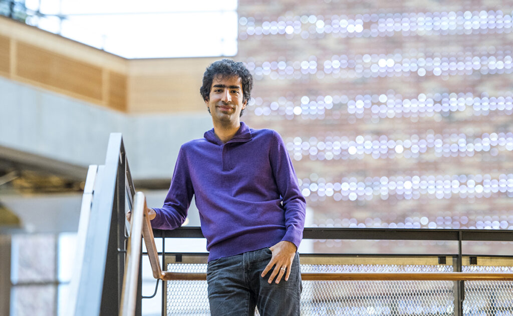 Shayan Oveis Gharan in a purple sweater and jeans leans against the wood and metal railing of the floating staircase in the atrium of the Paul G. Allen Center for Computer Science & Engineering. A wood and concrete balcony in front of floor-to-ceiling windows and a brick wall decorated in lights is blurred in the background.