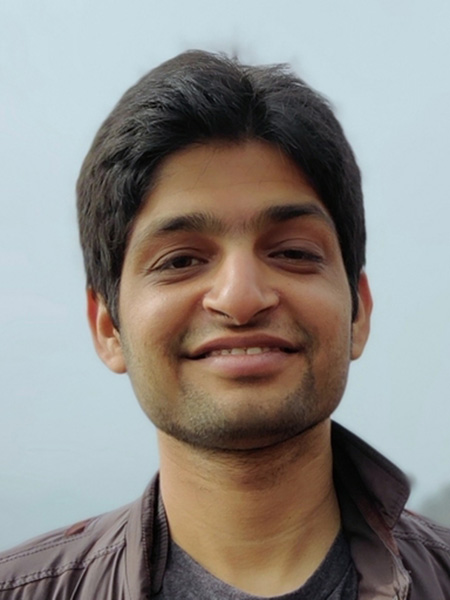 Ashish Sharma, wearing a brown shirt, smiles in front of a blurred blue background.