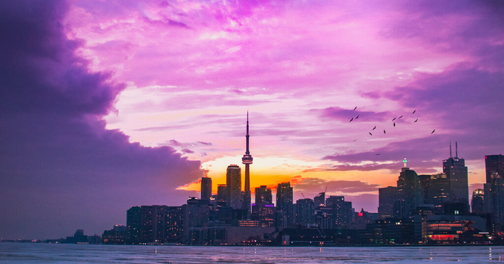 A nighttime view of Toronto. There is a pink and purple sky with clouds over the cityscape, and water in the foreground. The city is backlit from the setting sun, with the dark contours of the buildings visible. Dark outlines of birds are visible over the buildings on the right. 