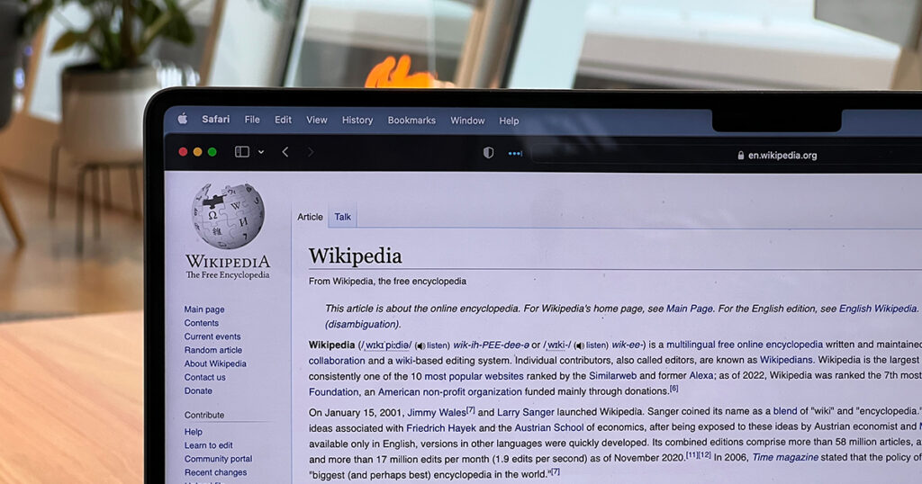 A photo shows a portion of a computer screen with the Wikipedia home page on the Safari browser. Behind the computer to the left is a table and the blurred background of a room with plants in pots on a stand and several windows behind it. 