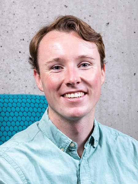 Portrait of Gus Smith wearing a pale aqua button-down shirt and seated in a teal upholstered chair against a textured concrete wall.