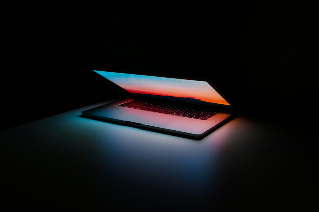 A partially open laptop with the screen illuminated in shades of blue, orange and red, which reflects off the keyboard and surrounding table. The laptop screen is the only source of light, with the background shrouded in darkness.