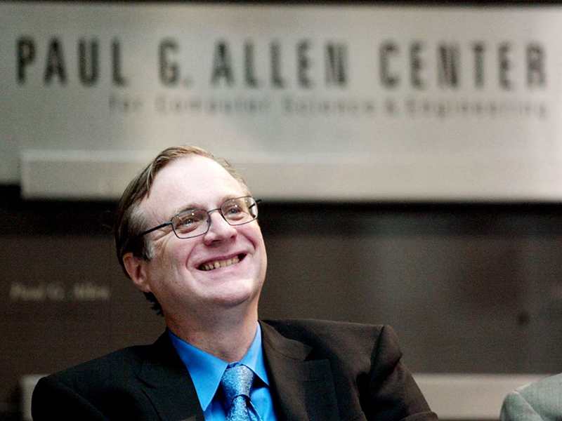 A smiling Paul Allen wearing glasses and a suit and tie seated in front of a metal sign displaying the building name, Paul G. Allen Center for Computer Science & Engineering