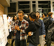 A crowd of people cluster around posters on easels on a building landing. The camera focuses on one person explaining a poster's contents to another person viewing it. The poster's content is not visible in the photo.