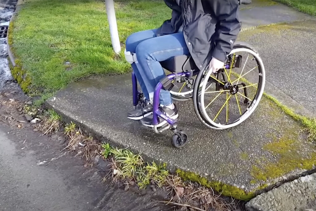 Person in wheelchair, pictured from the shoulders down, approaching the edge of a sidewalk at an intersection without a curb cut.