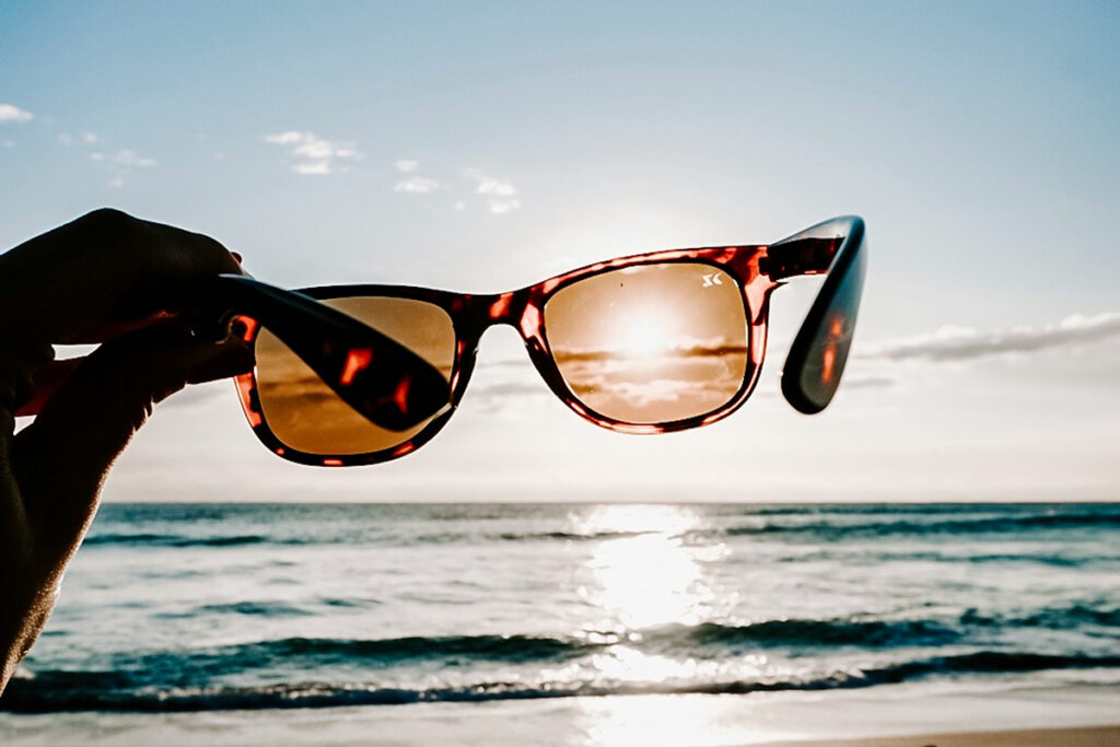 A person's hand is visible holding a pair of tortoiseshell sunglasses against the sky so that the sun shines through one lens while illuminating ocean waves at the shore