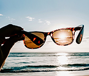 A person's hand is visible holding a pair of tortoiseshell sunglasses against the sky so that the sun shines through one lens while illuminating ocean waves at the shore