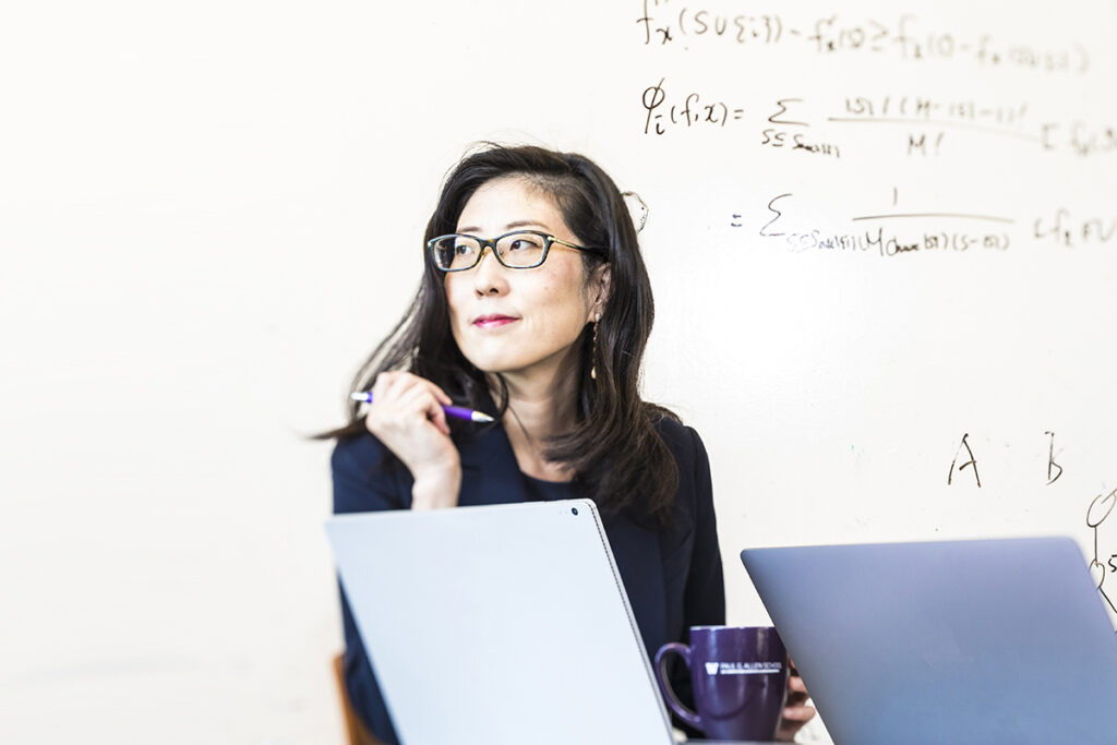 Su-In Lee looking off to the side with a slight smile on her face. She is holding a pen in one hand and an Allen School coffee mug in the other, seated behind an open laptop against the backdrop of a whiteboard with equations scribbled across it.