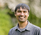 Shwetak Patel wearing a grey button-down shirt over a t-shirt smiling outdoors with foliage behind him.