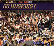Crowd of people seated in an arena underneath a sign saying Go Huskies! cheer for graduates seated on the arena floor below