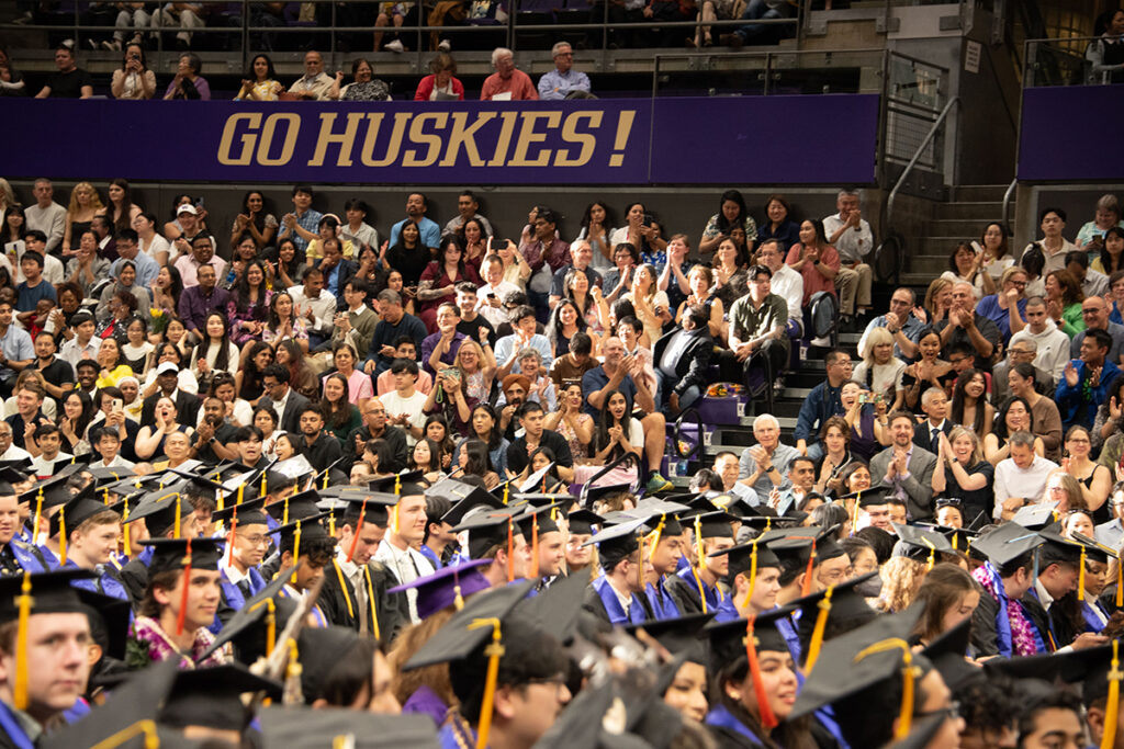 Crowd of people seated in an arena underneath a sign saying Go Huskies! cheer for graduates seated on the arena floor below