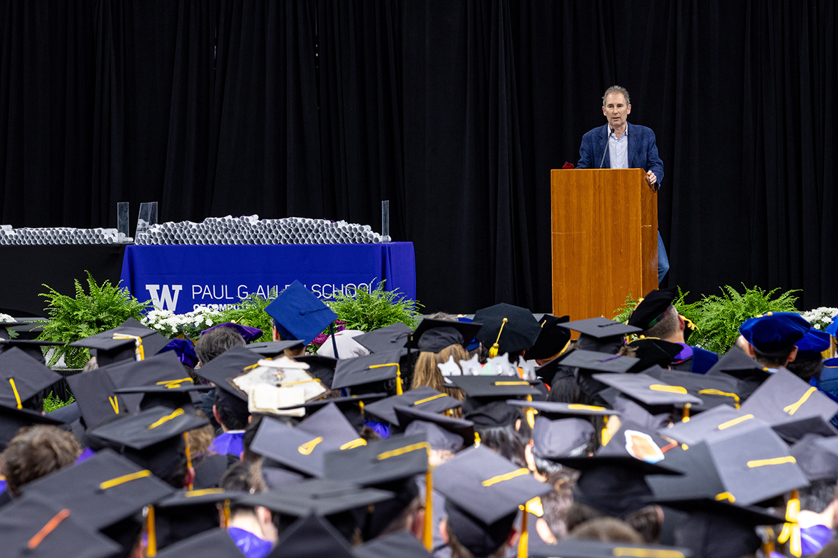 Andy Jassy address a crowd of graduates pictured in caps and gowns from a wooden podium onstage, with a table of souvenir diplomas off to the side