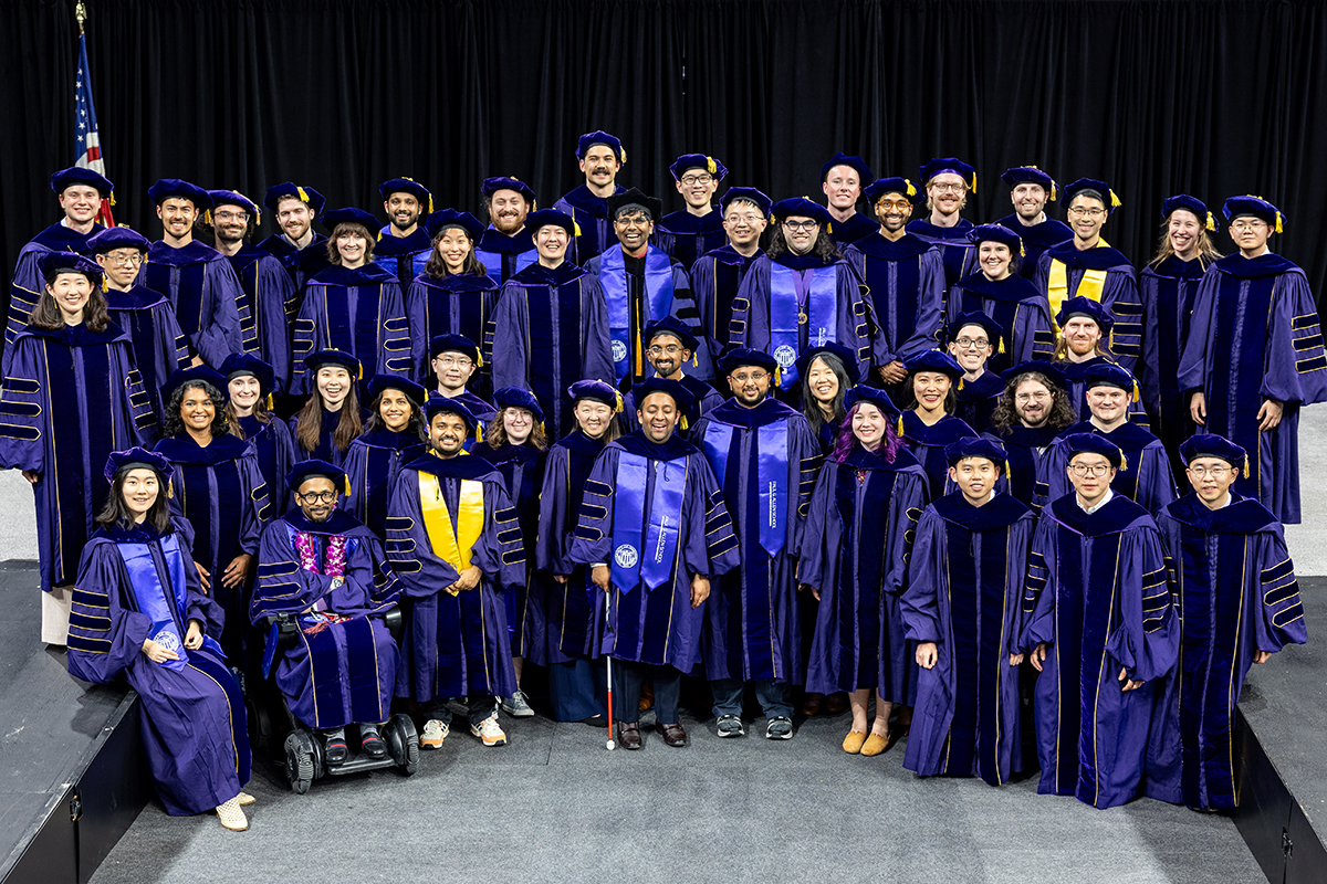 A group shot of 46 people dressed in doctoral regalia posing onstage