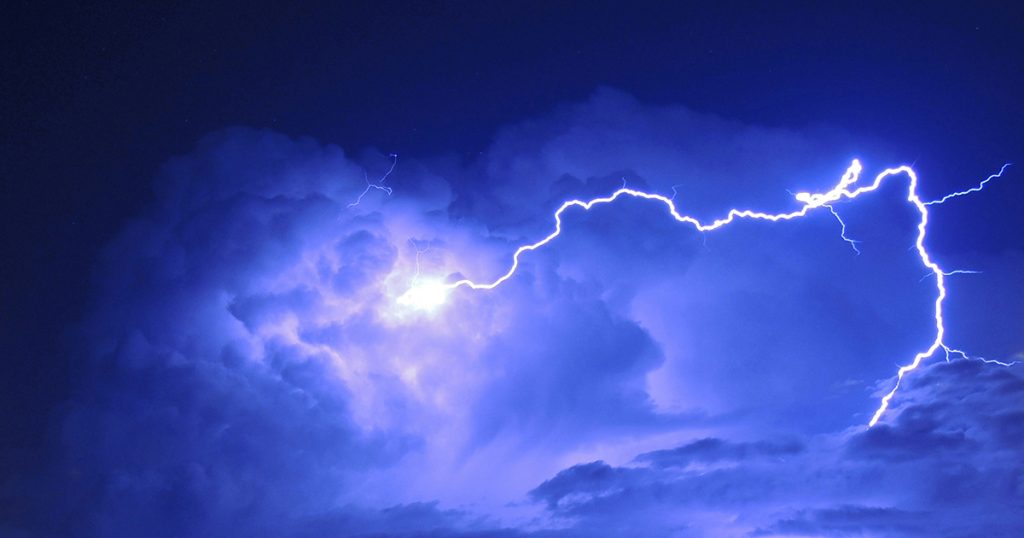 A large bolt of lightning illuminates clouds in a bluish-purple sky