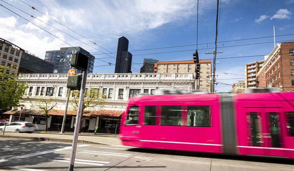 Urban street scene depicting hot pink streetcar entering an intersection, with a car on the opposite side of the road and painted pedestrian crossing visible against a backdrop of street trees and low-rise buildings, with high-rise buildings in distant background