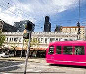 Urban street scene depicting hot pink streetcar entering an intersection, with a car on the opposite side of the road and painted pedestrian crossing visible against a backdrop of street trees and low-rise buildings, with high-rise buildings in distant background