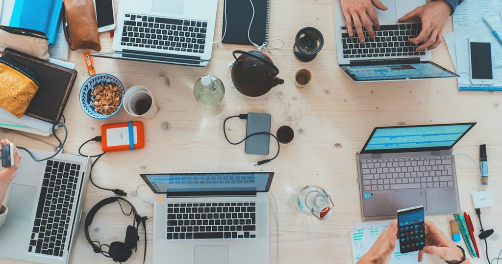 An overhead view of people sitting a table using laptops, phones, and headphones working on a project.