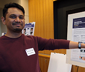 Anandghan Waghmare holds out his arm to display a smartwatch modified with additional, tiny sensors affixed to the armband in front of a poster on an easel