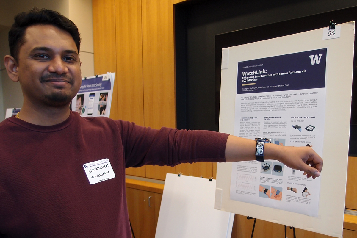 Anandghan Waghmare holds out his arm to display a smartwatch modified with additional, tiny sensors affixed to the armband in front of a poster on an easel titled WatchLink