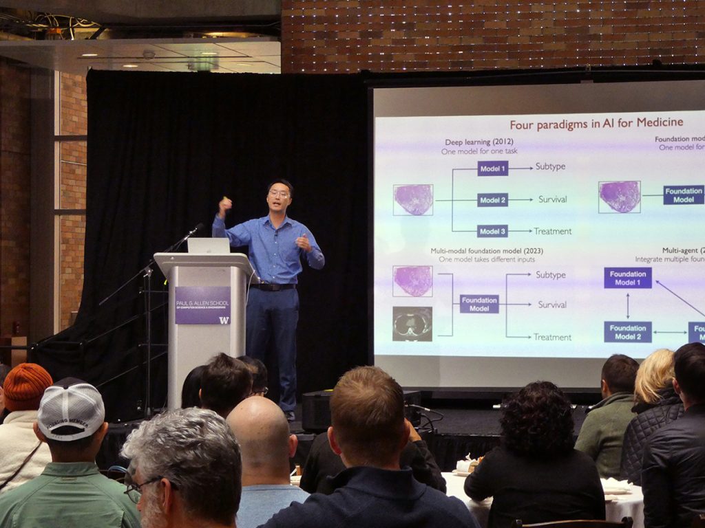 Sheng Wang onstage speaking to a crowd of people seated at round tables with a slide behind him titled Four Paradigms in AI for Medicine