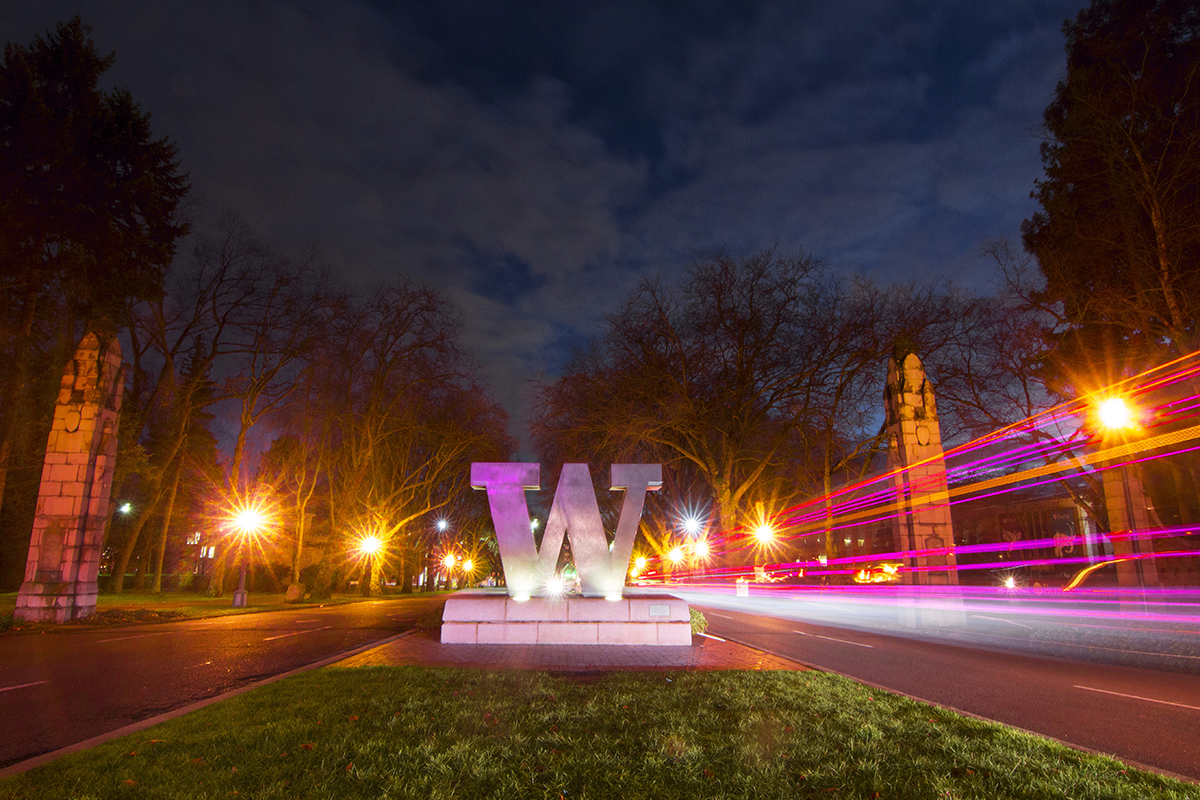 A bronze W statue at the entrance to the University of Washington campus at night, flanked by pink and orange tinged light trails from passing vehicles