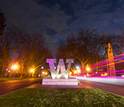 A bronze W statue at the entrance to the University of Washington campus at night, flanked by pink and orange tinged light trails from passing vehicles